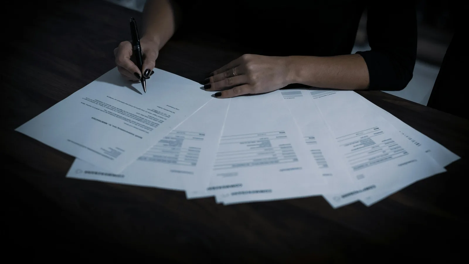 Mujer sentada a una mesa cubierta de papeles, firmando un documento con bolígrafo.